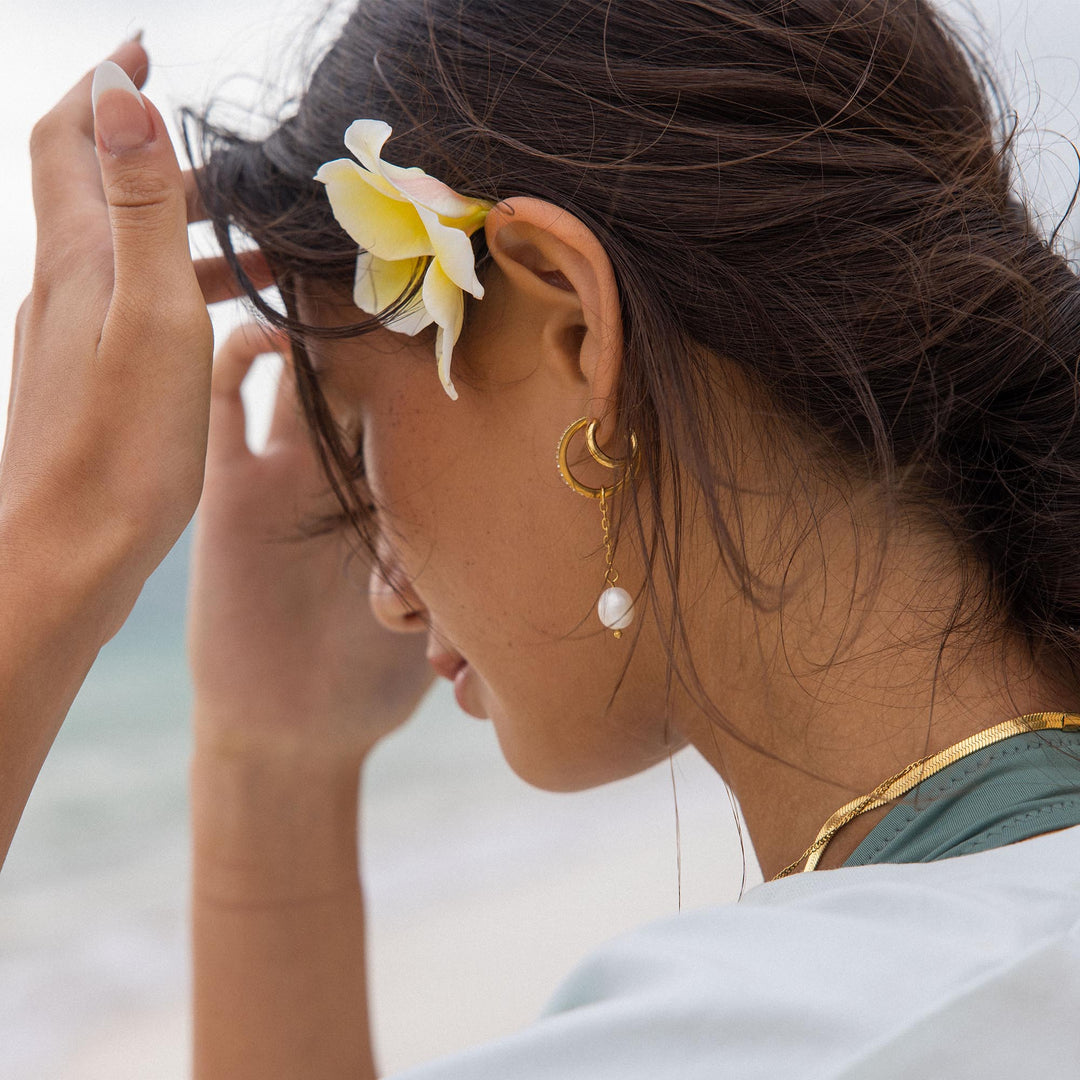 Gold double glitter earrings by PURELEI on a woman's left ear with a pearl-charm adding radiant sparkle to her style.