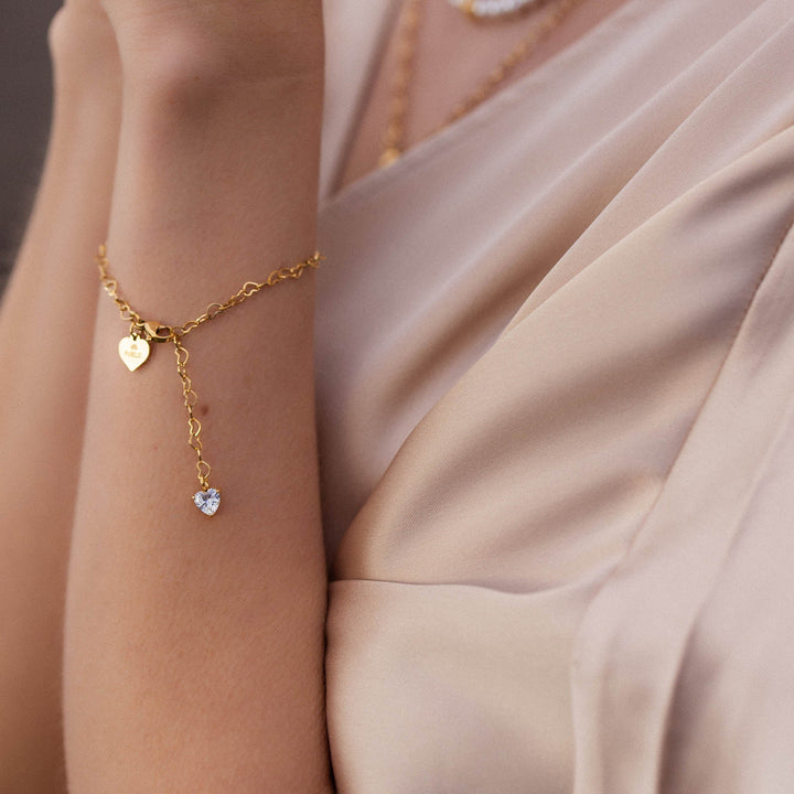 Woman wearing PURELEI Endless Love Bracelet on her left wrist, featuring gold heart-shaped links and a sparkling zirconia heart pendant.