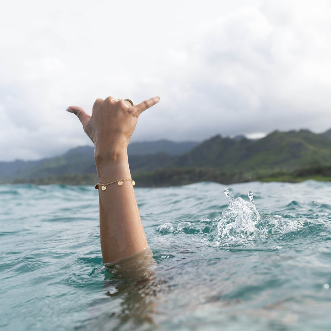 
A woman's right arm adorned with a gold kalea bracelet by PURELEI emerges from an infinity pool, with a stunning scenic background in view.