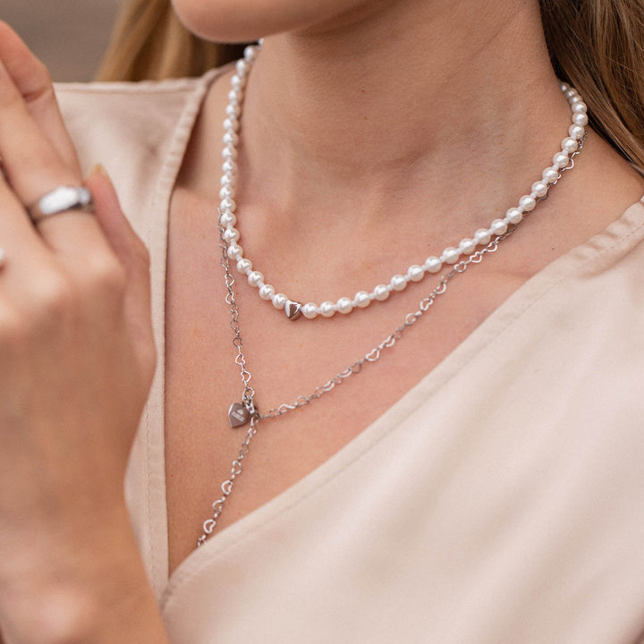 Woman layering the PURELEI Lovely Necklace in silver with another chain, showcasing the heart pendant and mixed pearls for a chic, elegant look.